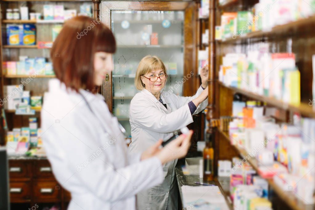 Young smiling female pharmacist in pharmacy reading the information about drugs. Middle-aged blond woman pharmacist choosing the drug on the background
