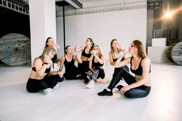 Beautiful fitness women in black clothes sitting on the floor and drinking water after fitness in the gym studio