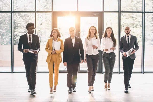 Business People Walking Toward Camera. Full length of group of happy young business people in formal wear having work meeting in modern office with large window