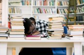 Etnické africká americká chlap spí u stolu uprostřed knihy v knihovně. Student je znuděnější.