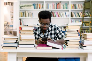 Koyu tenli öğrenci bilgi smartphone--dan okunurken üniversite kitaplığında okuyan, afro Amerikan hipster adam okuma mesaj mobil bankacılık online yapım hareketinde konsantre