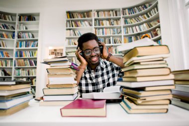 Kulaklıklar ile kütüphanede oturmuş ve bilimsel bir kitap okuma Afro-Amerikan öğrenci