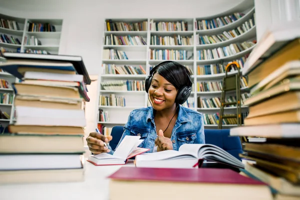 かなり若いアフリカの女性の座っていると彼女は近代的な大学図書館で彼女の本を勉強している間 音楽を聴きます 概念を勉強して — ストック写真