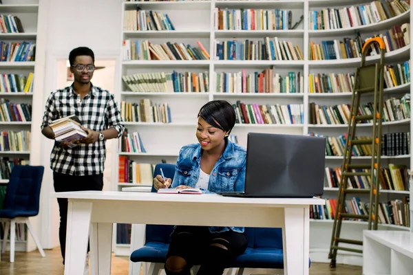 Pretty young african girl in casual clothes writing and studying with notebook and books in university library. Handsome african man in glasses holding books going to the girl on the background.