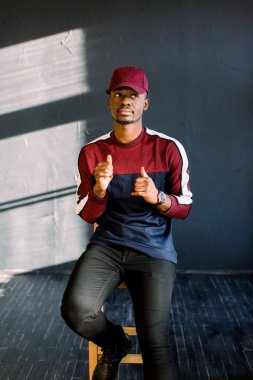 Mutlu genç Afrikalı Zenci bir tahta sandalyede karanlık bir arka plan üzerinde bir stüdyoda oturan kırmızı beysbol şapkalı portresi .