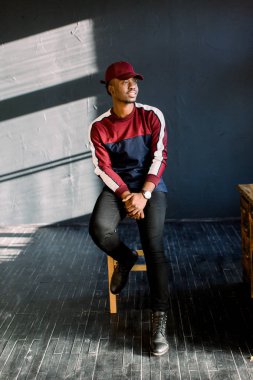 Bir Afrikalı-Amerikalı hipster adam portresi bir kırmızı yün kazak ve stüdyoda kırmızı şapka giymiş. Bir stüdyo ahşap sandalye üzerinde oturan karanlık dokulu arka plan üzerinde izole
