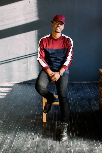 暗い背景に木の椅子でスタジオに座っている赤い野球帽の幸せな若い黒いアフリカ男の肖像 — ストック写真