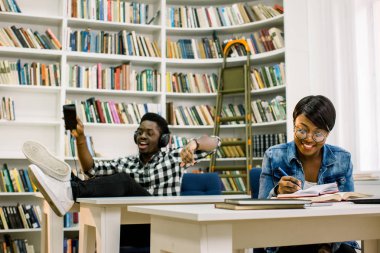 Güzel Afrikalı-Amerikalı öğrenciler kütüphanede çalışırlar. Kız gülümsüyor ve laptop ve çalışma, okuma kitapları ve çocuk dans müzik dinlerken.