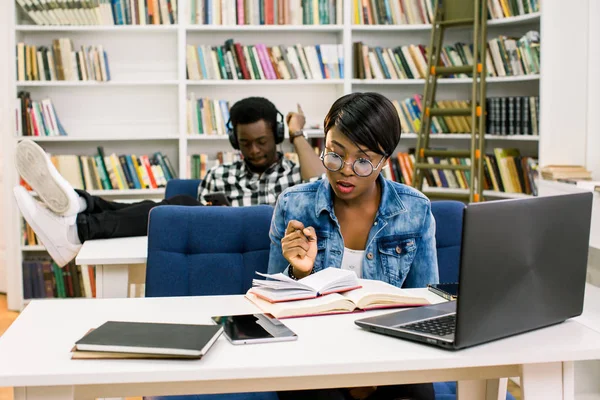図書館でアフロ アメリカ人学生 女の子は ラップトップを使用して 音楽に耳を傾け テーブルの上に彼の足を置く男の背景に本を読んでライブラリ — ストック写真