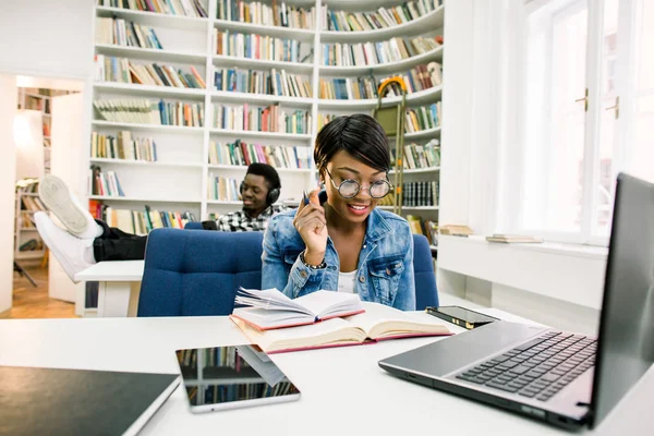 Happy afro-american student at university library sitting and laying legs on the table, listening to music and working with a smartphone. An African American girl prepares for exams in the library