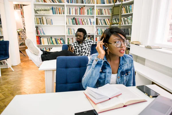 Happy afro-american student at university library sitting and laying legs on the table, listening to music and working with a smartphone. An African American girl prepares for exams in the library