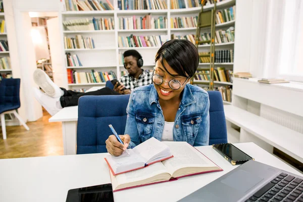 people, knowledge, education, literature and school concept - happy students reading books, listening to music, working with smartphone and preparing to exams in library