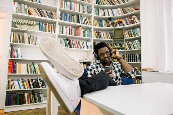 スマート フォンでの作業と音楽を聞いて大学図書館の机に座って美しい若いアフリカ学生 — ストック写真