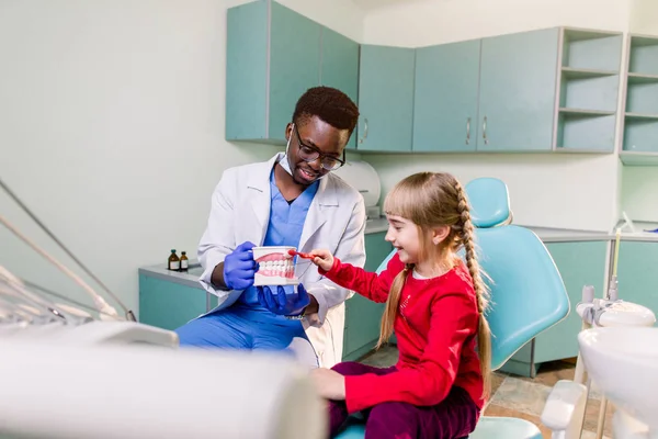 A smiling young African American dentist holds artificial jaws in his arms, and a little girl brushes with toothbrush teeth layout. Pediatric dentistry concept