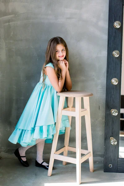 Happy beautiful surprised cute lovely charming small girl with toothy smile, she is wearing blue dress, put her hands on the stool, isolated on bright grey background, copyspace