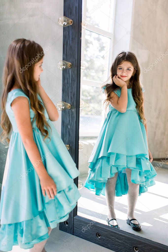 The lovely little girl posing in a beautiful blue dress is posing looking at the mirror