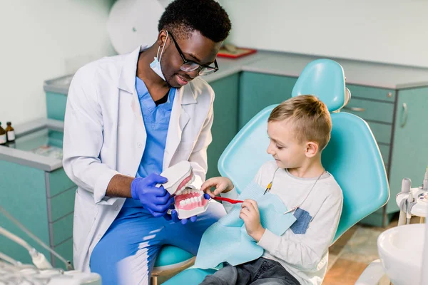 The child boy in dentist\'s chair having fun while brushing teeth with a male African American doctor