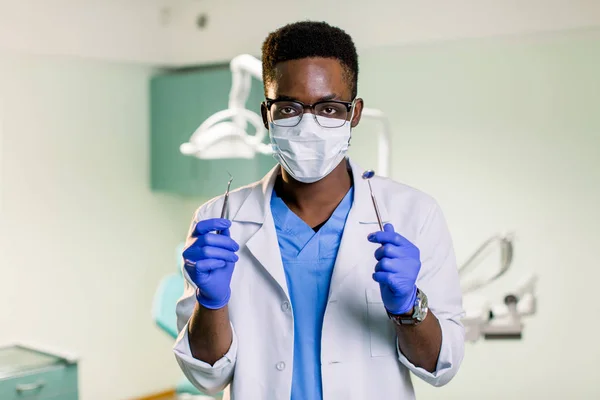Koruyucu yüz maskesi, beyaz önlük, mavi kravat, çekici yakışıklı siyah Dişçi aletleri onun kollarında tutan portresi