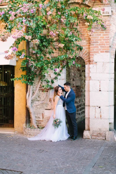 A beautiful young couple in a wedding lothes poses against a background of a wall of an old house with a flowering tree. Sirmione, Italy