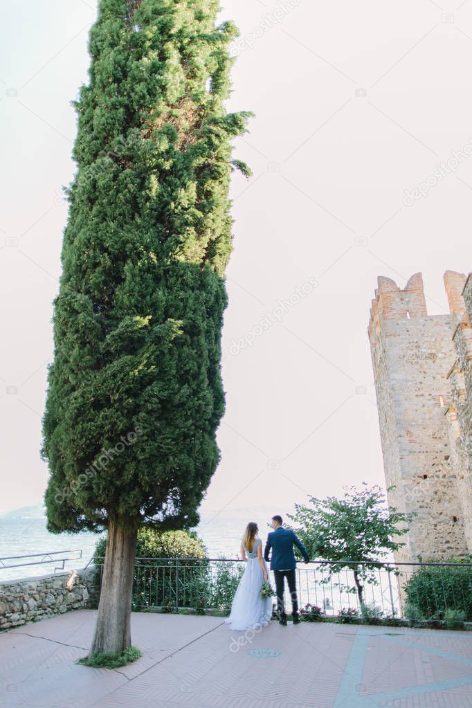 Beautiful young woman in blue dress and handsome man in suit walking in the garden of Rocca Scaligera medieval castle, Sirmione town, Garda lake, Italy