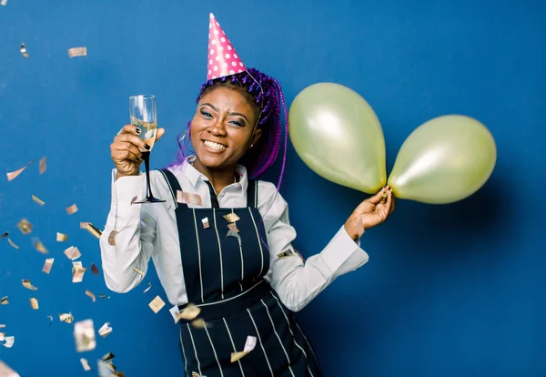 Portrait of beautiful african girl in pink party cap holding air balloons and champagne, resting at party. African woman having fun at the party with confetti on blue background