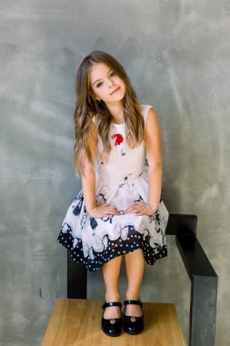Beautiful little girl with long broun hair and blue eyes sitting on the chair on a grey background wearing a black and white spring dress clipart