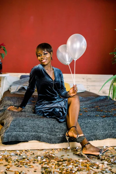 Happy dark-skinned girl in a dark evening dress is lying on the bed after the birthday party.