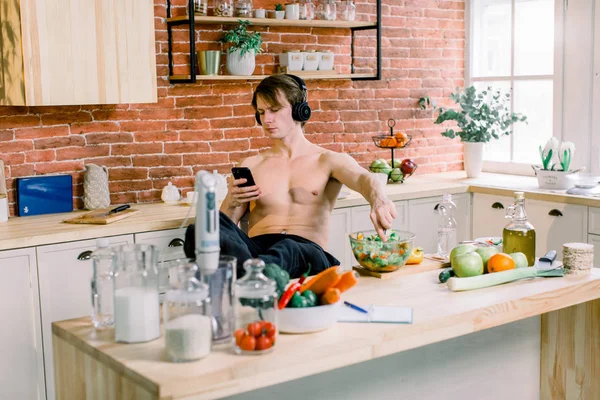 Healthy and vegan food concept. Happy Caucasian young man in earphones listening to music and eating carrot and making in salad from fresh vegetables in the modern kitchen