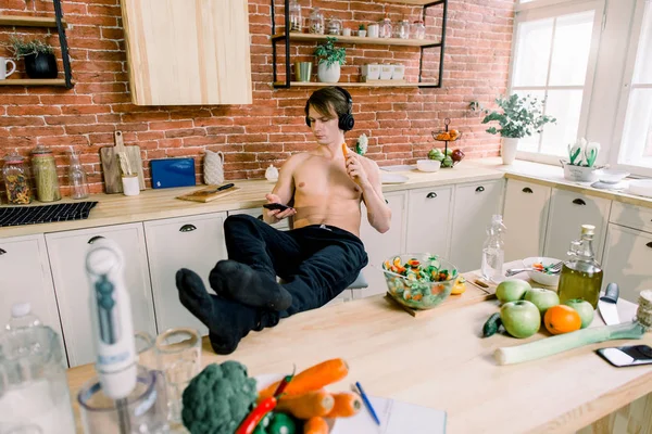 帅哥在家里做饭 在厨房里准备沙拉 年轻人正在听耳机里的音乐 在准备沙拉的时候使用智能手机和吃胡萝卜 健康的生活方式和食物 — 图库照片
