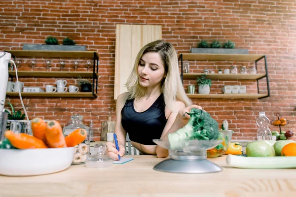 Young Woman Cooking Vegetable Salad and weights broccoli while sitting in the kitchen on the background of red brick wall. Dieting Concept. Healthy food and Lifestyle. Cooking At Home.