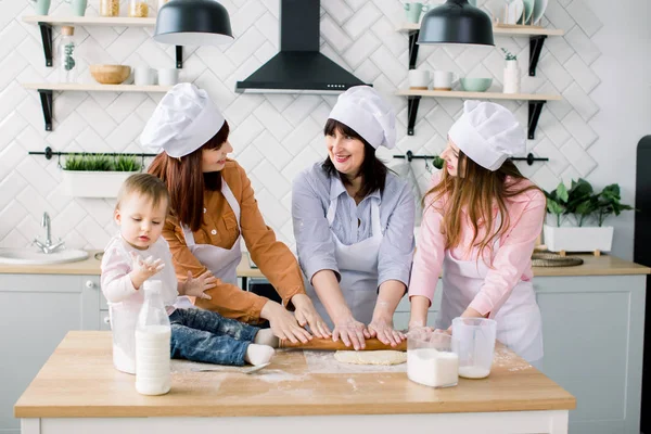 Smiling middle aged woman in kitchen apron rolling out dough and two daughters helping her. Litle baby girl sitting on the table and having fun. Happy women in white aprons baking together