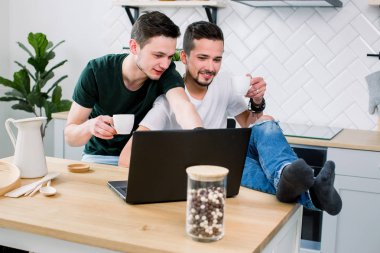 Tarafından mutfak ev dizüstü bilgisayar önünde masada oturan ve online haber izlerken çağdaş Eşcinsel bir çift. Kot pantolon ve t-shirt mutlu gay çift birlikte vakit