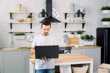 Beyaz t-shirt genç yakışıklı erkekler evde modern mutfak ahşap masa yanında dururken dizüstü kullanarak