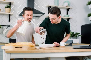 Sabah mutfakta kahvaltı hazırlarken mutlu gay çift. Yakışıklı adam ortağı kasenin içine süt dökme sırasında beyaz kase kahvaltılık koyarak