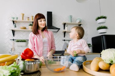 Genç güzel anne modern ışık mutfakta taze salata için yeşil salata doğrama küçük bebek kızı arıyorum. Birlikte, sağlıklı beslenme, annelik pişirme