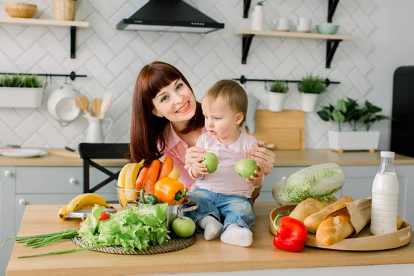 Anya és kis gyerek, egészséges étel és a szórakozás. Kislány ül a fa asztal, friss zöldségek és gyümölcsök, és tart alma — Stock Fotó
