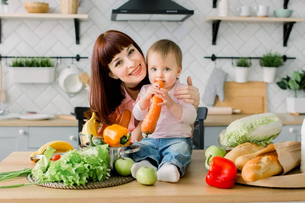 Feliz rindo menina da criança comendo a cenoura e sua bela jovem mãe pronta para preparar salada fresca para o café da manhã juntos em uma cozinha de luz ensolarada — Fotografia de Stock