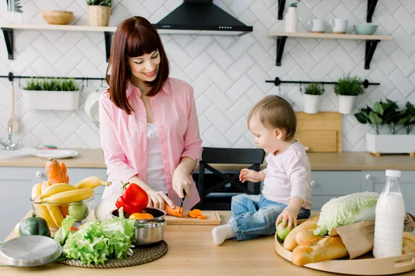 Mosolygó fiatal anya Faaprító sárgarépa felkészülése friss salátával és a kis aranyos baba kisgyermek gazdaság zöld alma és ül az asztalon, konyhában. Baba segít anya főzni, az otthoni konyha — Stock Fotó