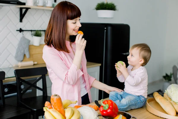 Mãe feliz e pequena menina se divertindo enquanto cozinham juntos na cozinha. Mãe comendo cenoura e bebê comendo maçã verde. Estilo de vida saudável, comida fresca, culinária familiar — Fotografia de Stock