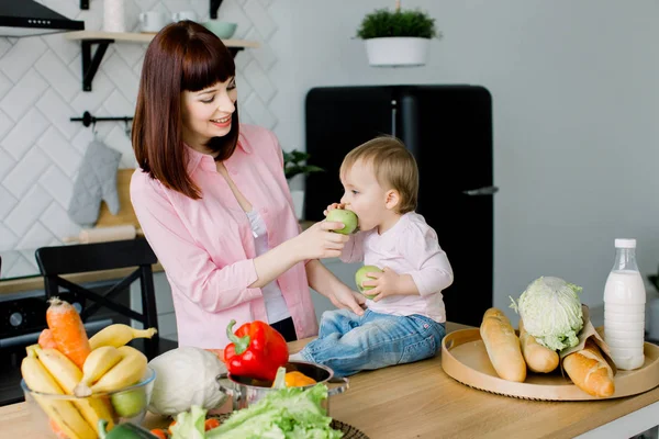 Fiatal anya reggelijét mosolyogva és etetés neki kis baby lánya zöld alma konyha — Stock Fotó