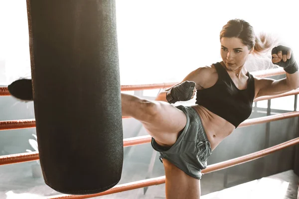 Jovem treina no ringue de boxe com saco de perfuração pesado. Retrato de um modelo de moda sexy treino no loft ginásio vintage. Pontapés nos pés ou no joelho . — Fotografia de Stock