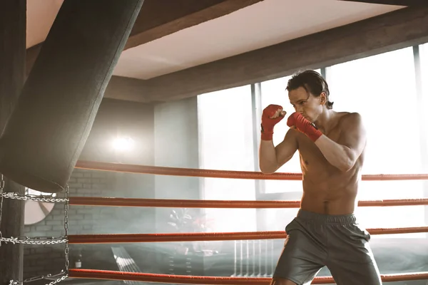 Treinamento de boxeador masculino com saco de perfuração no salão de esportes escuro. Treinamento de pugilista jovem em saco de perfuração. Boxer masculino como exercício para a grande luta. Boxer bate saco de perfuração. — Fotografia de Stock