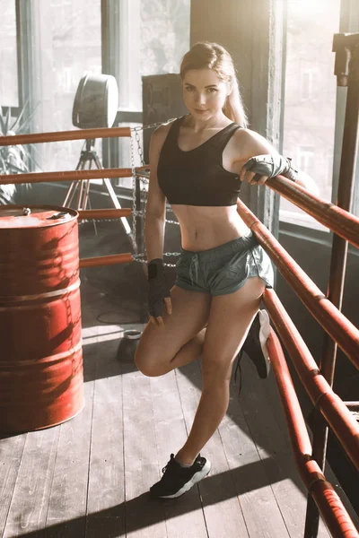 Young pretty boxer woman standing on ring. Full body portrait of boxer woman wearing black sports bra, grey trousers, trainers standing in ring and leaning on ropes. Boxing, sport concept