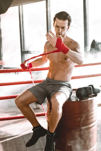 Jonge knappe bokser trekt pleister voor de strijd of de training zittend op de rode vat in boksring. Boksen Fighter Bandages voorbereiden opleiding — Stockfoto