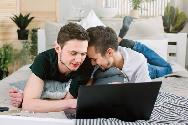 Привлекательный молодой человек опирается на своего парня в постели, наблюдает за ноутбуком. Пара геев лежит на кровати и использует ноутбук дома
