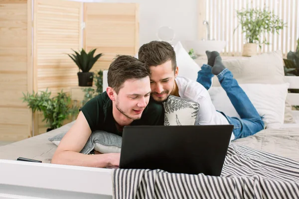 Feliz pareja gay acostada en la cama con el ordenador portátil en casa —  Fotos de Stock