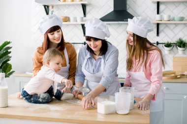 Annesi, teyzesi ve büyükannesi çerez hamur yaparken küçük bebeği Ahşap mutfak masasında oturuyor. Beyaz önlük birlikte pişirme mutlu kadın