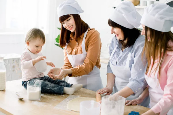 Little baby girl is sitting on the wooden table at kitchen and having fun with sugar. Grandmother and her daughters are baking cookies. Happy women in white aprons baking together. Mothers Day — Stock Photo, Image