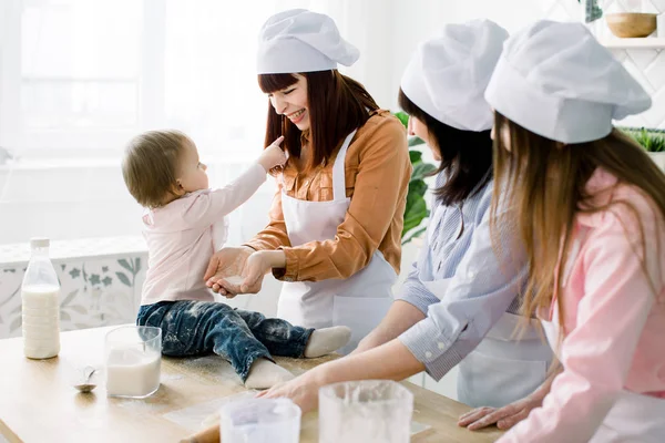 Little baby girl is sitting on the wooden table at kitchen and having fun with sugar. Grandmother and her daughters are baking cookies. Happy women in white aprons baking together. Mothers Day — Stock Photo, Image