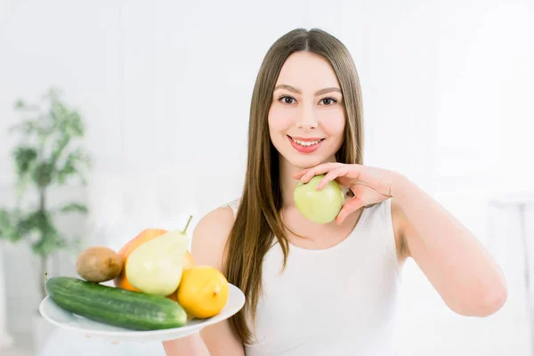 Υγιές, όμορφο χαμόγελο, χαριτωμένο κορίτσι που χαμογελά. Πορτρέτο ενός κοριτσιού με πιάτο φρούτα στο ένα χέρι και ένα μήλο σε ένα άλλο. — Φωτογραφία Αρχείου